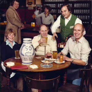 Gruppenfoto am Stammtisch mit großem Bembel „50 Jahre Wagner“, Menschen mit Apfelwein und Speisen.